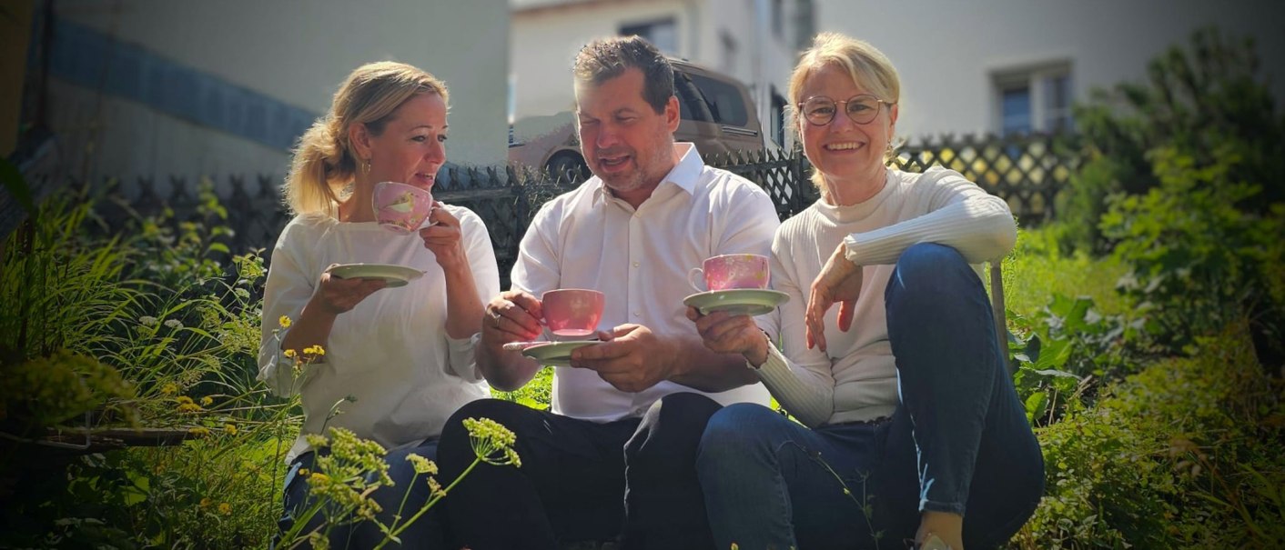 From left to right: Annette Ehrlich, Holger Köckritz and Annette Currle sit in the garden and enjoy tea from pink cups. They are wearing white tops and smiling at the camera., © Perle Uhlbach From left to right: Annette Ehrlich, Holger Köckritz and Annette Currle sit in the garden and enjoy tea from pink cups. They are wearing white tops and smiling at the camera., © Perle Uhlbach