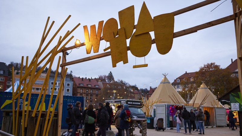 Eingang zum Wouahou Winterdorf mit gelbem Schild, Menschen und Tipis im Hintergrund. Es ist Abend und die Beleuchtung ist eingeschaltet., © SMG, Sarah Schmid