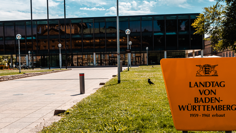 The state parliament of Baden-Württemberg with a modern glass façade and a yellow sign in the foreground. The television tower can be seen in the background., © Stuttgart-Marketing GmbH, Sarah Schmid The state parliament of Baden-Württemberg with a modern glass façade and a yellow sign in the foreground. The television tower can be seen in the background., © Stuttgart-Marketing GmbH, Sarah Schmid