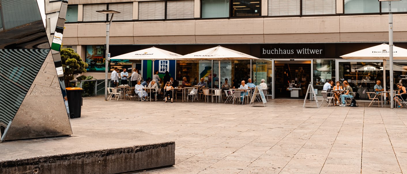 Menschen sitzen unter Sonnenschirmen vor einem Café neben dem Buchhaus Wittwer. Im Hintergrund ist ein Fitnessstudio zu sehen., © SMG, Sarah Schmid Menschen sitzen unter Sonnenschirmen vor einem Café neben dem Buchhaus Wittwer. Im Hintergrund ist ein Fitnessstudio zu sehen., © SMG, Sarah Schmid