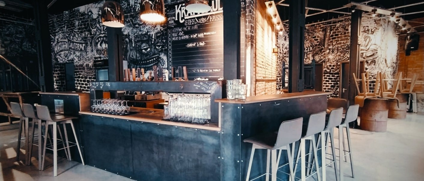 Moderne Bar mit industriellem Design, hohen Stühlen und Wandkunst. Gläser stehen auf der Theke, im Hintergrund eine Tafel mit Getränken., © Kraftpaule