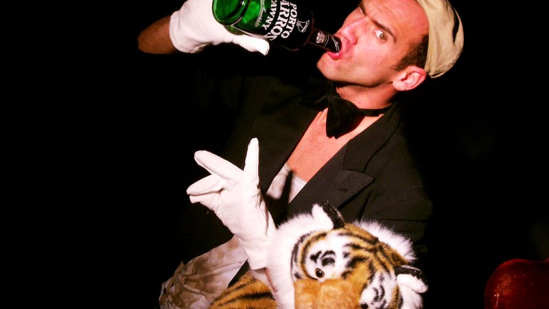 Ein Mann in einem Kostüm trinkt aus einer Flasche und hält einen Plüschtiger. Er trägt weiße Handschuhe und eine Fliege., © Nelly Eichhorn Ein Mann in einem Kostüm trinkt aus einer Flasche und hält einen Plüschtiger. Er trägt weiße Handschuhe und eine Fliege., © Nelly Eichhorn