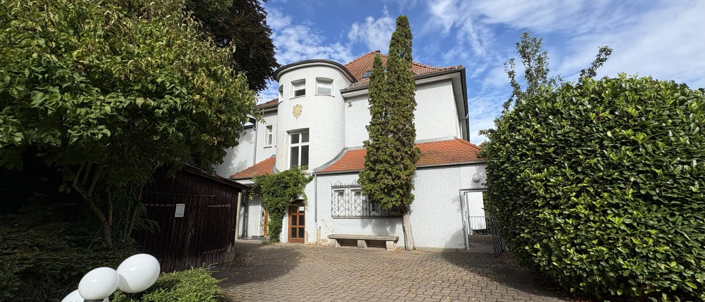 Ein weißes Haus mit rotem Dach und rundem Turm, umgeben von Bäumen und Sträuchern. Der gepflasterte Hof führt zum Eingang., © TOMAS Ein weißes Haus mit rotem Dach und rundem Turm, umgeben von Bäumen und Sträuchern. Der gepflasterte Hof führt zum Eingang., © TOMAS