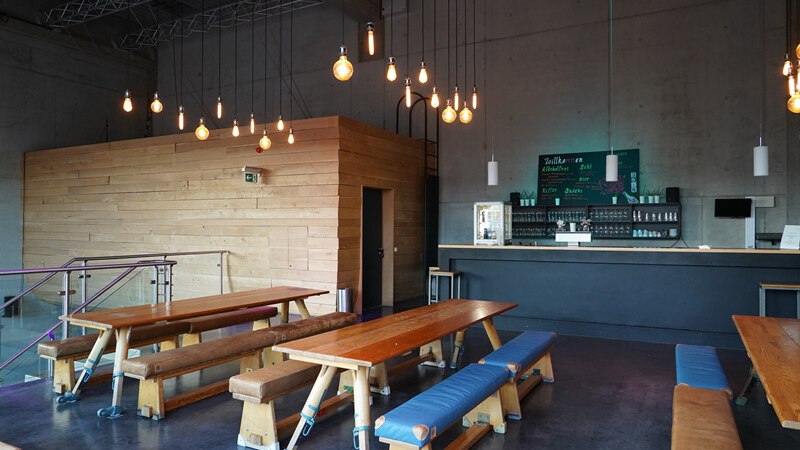 Modernes Caf&eacute; mit Holztischen und B&auml;nken, H&auml;ngelampen und einer Bar im Hintergrund. Die W&auml;nde sind aus Beton, der Raum wirkt industriell., &copy; W&uuml;rttembergische Staatstheater Stuttgart