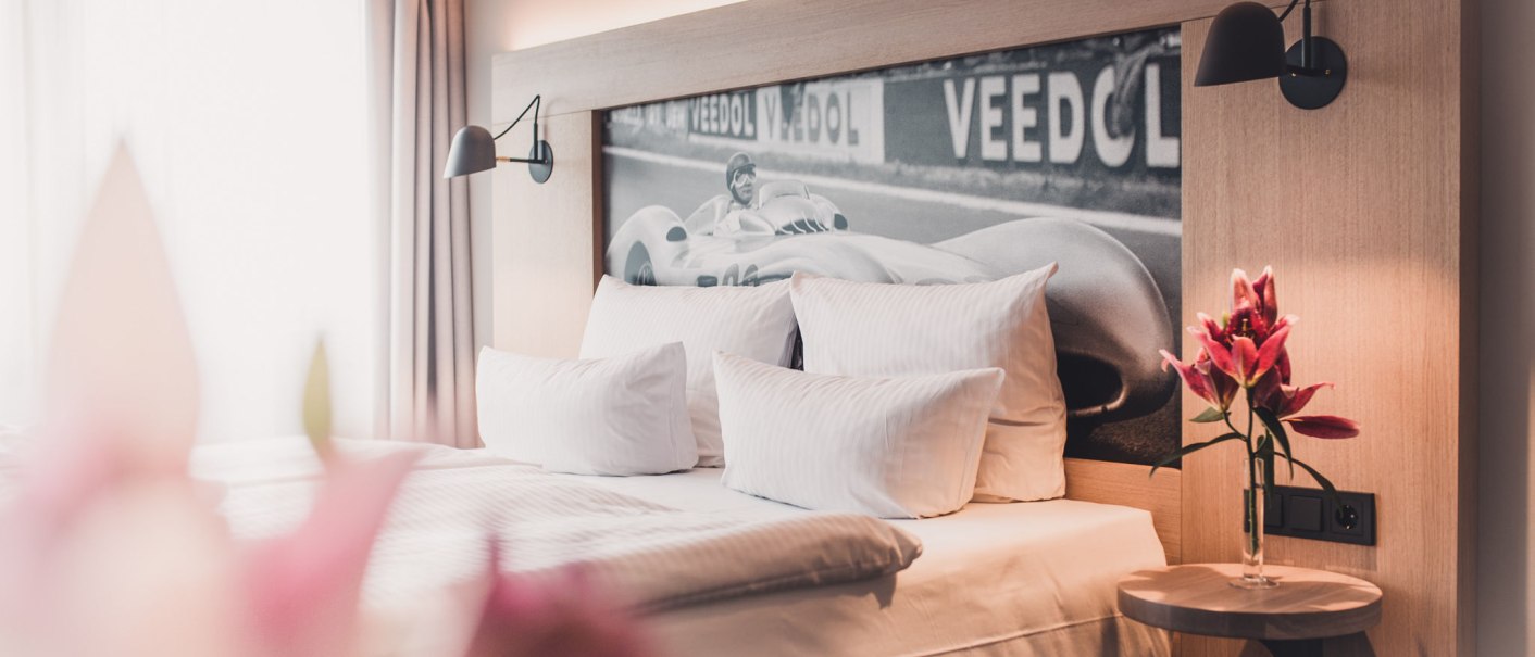 Hotelzimmer mit Doppelbett, stilvollem Rennwagenbild im Hintergrund und Blumen auf einem kleinen Tisch., © Abacco Hotels GmbH Hotelzimmer mit Doppelbett, stilvollem Rennwagenbild im Hintergrund und Blumen auf einem kleinen Tisch., © Abacco Hotels GmbH