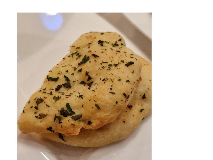 Zwei St&uuml;cke indisches Fladenbrot, bestreut mit Kr&auml;utern, liegen auf einem wei&szlig;en Teller. Das Brot wirkt frisch und aromatisch., &copy; Kultweine-Kochwelten-Kunst&uuml;cke - Kochschule Sonja Lenz