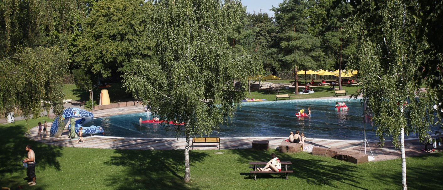Ein Freibad mit grüner Liegewiese, Bäumen und Schwimmern im Wasser. Menschen entspannen auf Bänken und unter gelben Sonnenschirmen., © Stuttgarter Bäder