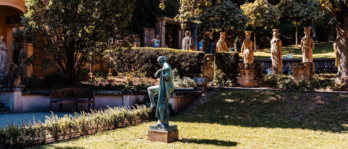 Ein sonniger Garten mit antiken Statuen, umgeben von Bäumen und Sträuchern. Eine Bank steht im Schatten, während eine Statue im Vordergrund hervorsticht., © Stuttgart Marketing GmbH, Sarah Schmid