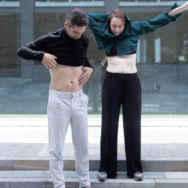 Zwei Personen heben ihre Oberteile und zeigen ihre Bäuche. Sie stehen vor einem modernen Gebäude mit Glasfassade., © Renitenztheater Stuttgart e.V.