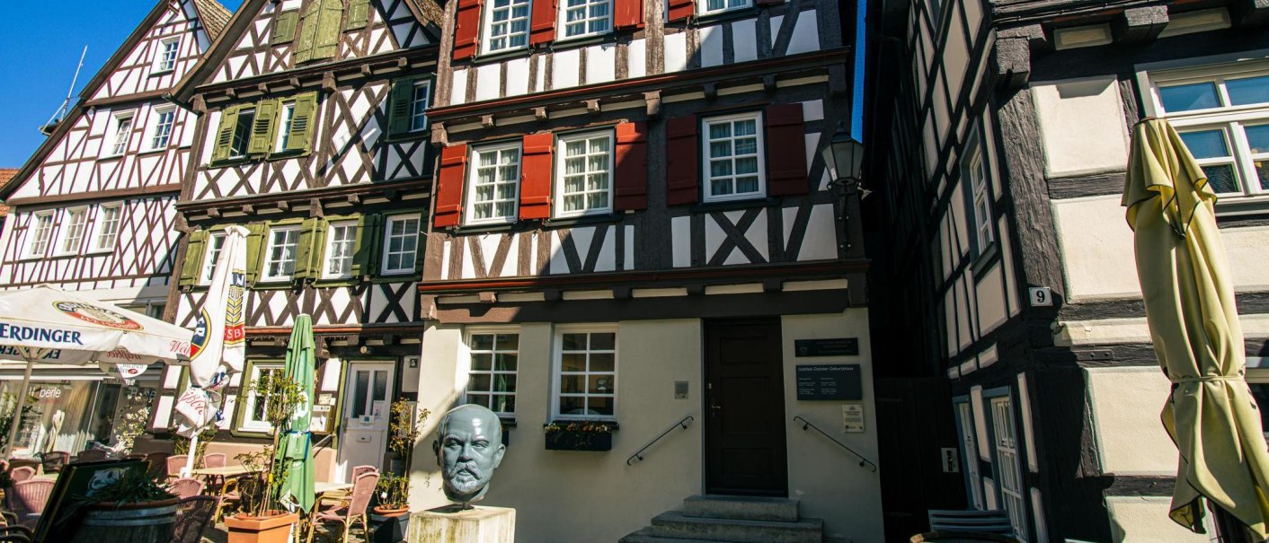 Fachwerkh&auml;user in Schorndorf mit einer B&uuml;ste von Gottlieb Daimler im Vordergrund. Sonnenschirme und ein Caf&eacute;bereich sind ebenfalls zu sehen., &copy; Stuttgart-Marketing GmbH, Sarah Schmid