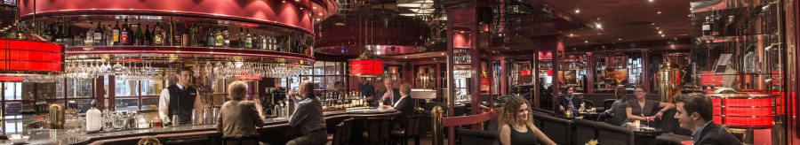 Elegant bar with red d&eacute;cor, mirrors and subdued lighting. Guests sit at the counter and at tables while a bartender serves drinks., &copy; Maritim Hotels