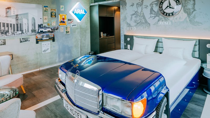 Ein Hotelzimmer im V8 Hotel mit einem Bett in Form eines blauen Mercedes-Autos, Wanddekorationen und einem ARAL-Schild., © Thomas Niedermüller Ein Hotelzimmer im V8 Hotel mit einem Bett in Form eines blauen Mercedes-Autos, Wanddekorationen und einem ARAL-Schild., © Thomas Niedermüller
