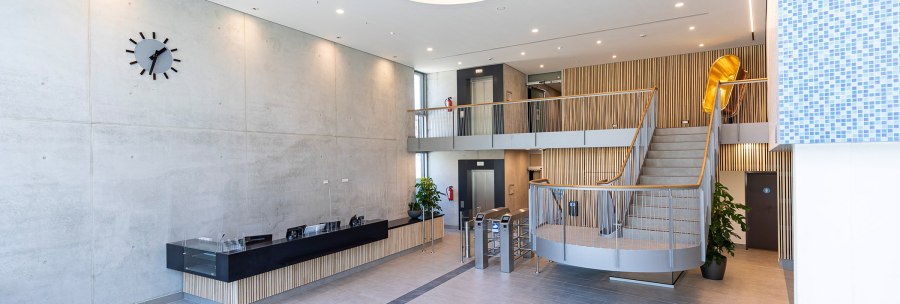 Modern foyer with reception desk, staircase, elevators and a large wall clock. The ceiling has a colorful glass element., © Stuttgarter Bäder