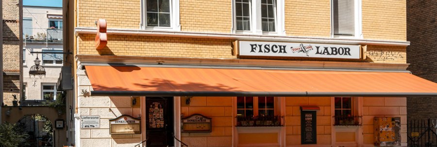 A restaurant called Fischlabor with an orange awning and outdoor seating. The building has yellow brick walls and white window frames., © SMG Stuttgart Marketing GmbH - Sarah Schmid