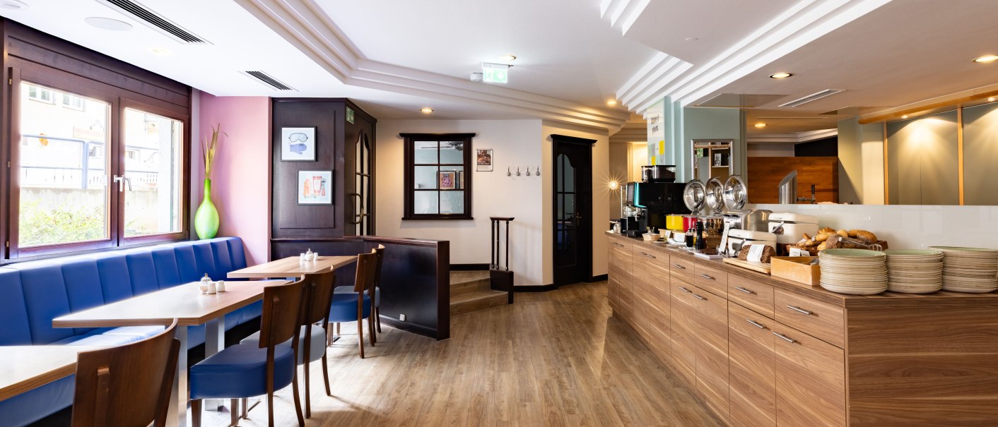 A bright breakfast room with blue benches, wooden floors, and a buffet with dishes and food. Large windows let in natural light., &copy; Novum  Hospitality