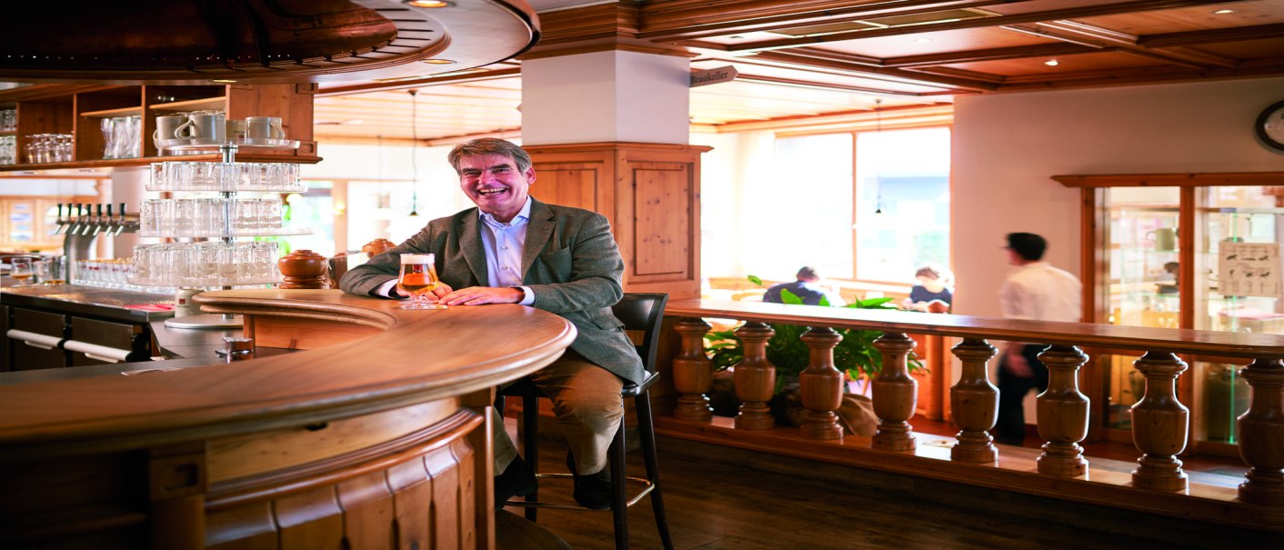 A man sits smiling at a wooden bar in a brewery pub, holding a glass of beer. Guests and a waiter can be seen in the background., © © Stuttgart-Marketing GmbH A man sits smiling at a wooden bar in a brewery pub, holding a glass of beer. Guests and a waiter can be seen in the background., © © Stuttgart-Marketing GmbH
