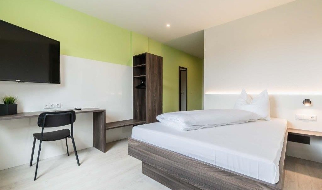 A modern single room with a bed, desk, chair, and TV. The walls are painted green and white, and the furniture is made of dark wood., &copy; a2 HOTELS am Quadrium