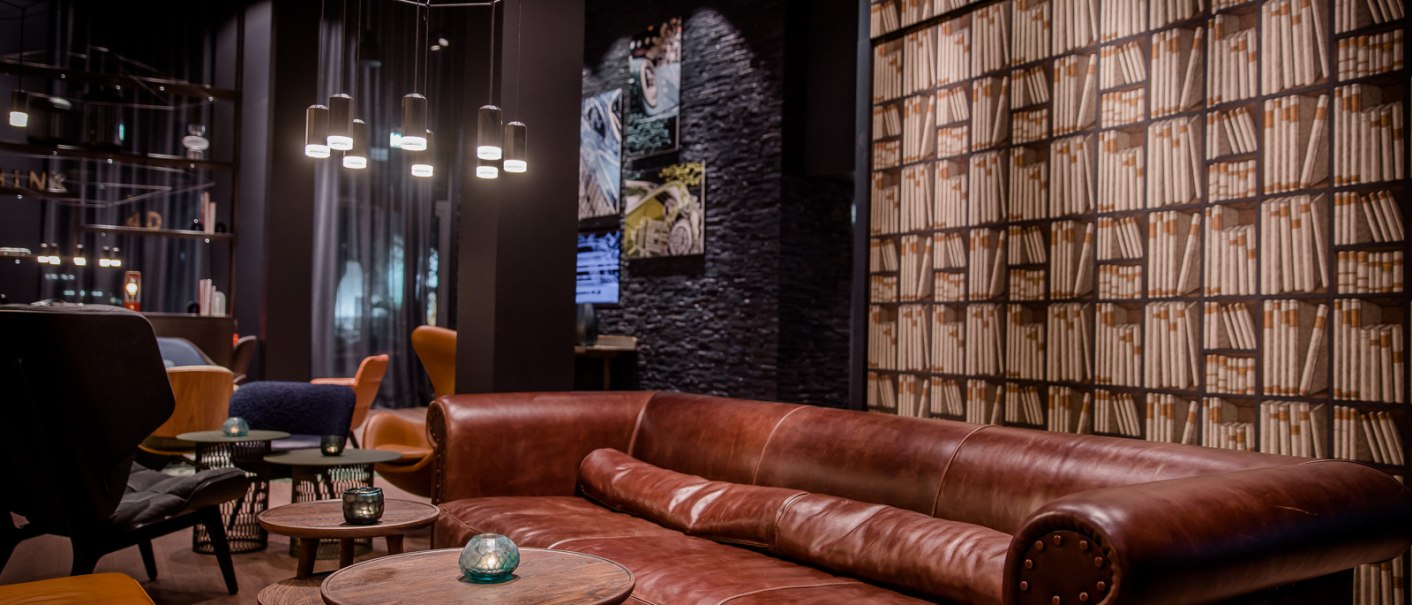 Eleganter Lounge-Bereich mit braunem Ledersofa, runden Holztischen und Bücherregal-Tapete. Moderne Beleuchtung und gemütliche Atmosphäre., © TOMAS