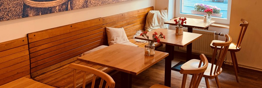 Cozy interior of a café with wooden tables and chairs, flower vases on the tables and a large window with a view outside., © Gustav Gastro GmbH Cozy interior of a café with wooden tables and chairs, flower vases on the tables and a large window with a view outside., © Gustav Gastro GmbH