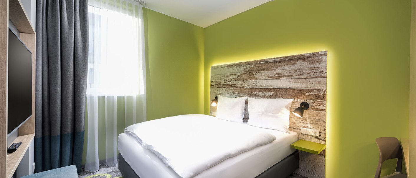 Modernes Doppelzimmer mit grün gestrichener Wand, großem Bett, Fernseher und Fenster mit Vorhängen., © ibis Styles Stuttgart Vaihingen