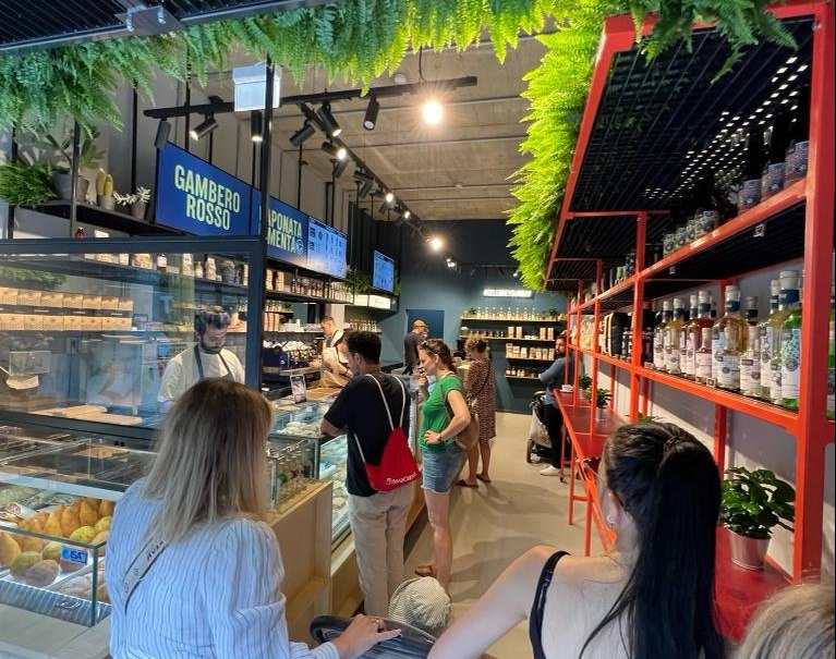 Lively delicatessen with plants on the ceiling, customers in a queue, shelves with products and a counter with food., © CELEBRE