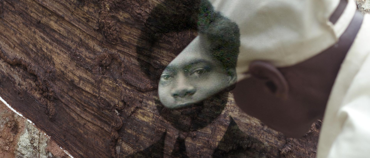 Ein Mann in weißer Kleidung betrachtet ein altes Foto, das auf die Rinde eines Baumes projiziert ist. Das Bild zeigt ein Gesicht., © Twenty Nine Studio