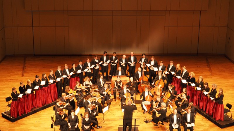 Ein Chor und Orchester auf einer Bühne. Der Dirigent steht vor den Musikern, die in formeller Kleidung auftreten. Der Konzertsaal ist gut beleuchtet., © Musik Podium Stuttgart