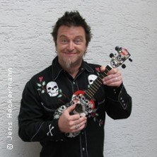 Ein Mann in einem schwarzen Hemd mit Totenkopf-Motiven hält eine Ukulele und lächelt in die Kamera. Hintergrund ist eine weiße Wand., © links im Bild