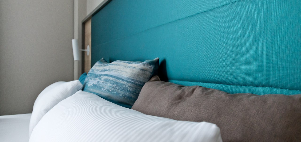 Nahaufnahme eines Bettes mit t&uuml;rkisfarbenem Kopfteil und Kissen in Wei&szlig;, Blau und Braun. Modernes, gem&uuml;tliches Design., &copy; Arthotel Ana / elaya
