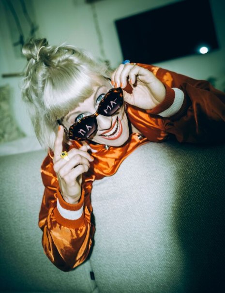 Eine fröhliche Frau mit blonden Haaren trägt eine Sonnenbrille mit der Aufschrift "Mama" und eine orangefarbene Jacke., © Theaterhaus Stuttgart e.V. Eine fröhliche Frau mit blonden Haaren trägt eine Sonnenbrille mit der Aufschrift "Mama" und eine orangefarbene Jacke., © Theaterhaus Stuttgart e.V.