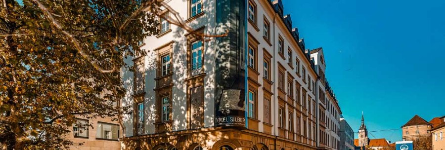 The Hotel Silber, a historic building, stands on a sunny street. Trees cast shadows on the façade and a church is visible in the background., © Haus der Geschichte Baden-Württemberg
