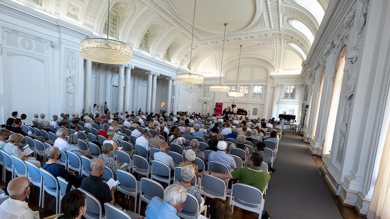 Ein eleganter, wei&szlig;er Saal mit hohen Decken und Kronleuchtern. Viele Menschen sitzen auf St&uuml;hlen und schauen nach vorne, wo ein Konzert stattfindet., &copy; Internationale Hugo-Wolf-Akademie