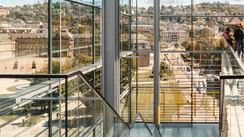 Blick durch moderne Glasarchitektur auf den Schlossplatz. Im Hintergrund historische Gebäude und Menschen. Klare Linien und Reflexionen dominieren das Bild., © Stuttgart-Marketing GmbH, Sarah Schmid Blick durch moderne Glasarchitektur auf den Schlossplatz. Im Hintergrund historische Gebäude und Menschen. Klare Linien und Reflexionen dominieren das Bild., © Stuttgart-Marketing GmbH, Sarah Schmid