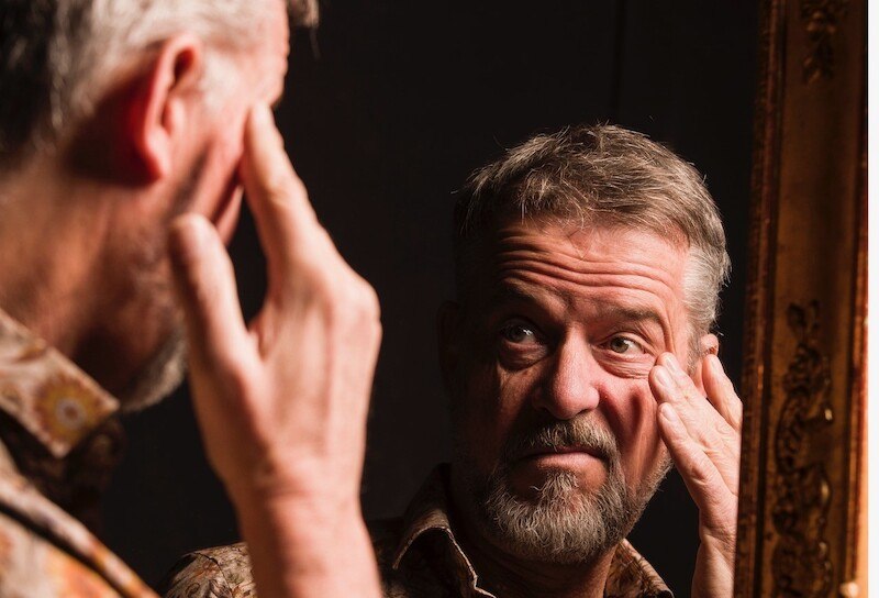A man with a beard looks thoughtfully into a mirror and touches his face. The background is dark, the frame of the mirror is decorative., &copy; Rosenau Kultur e.V.