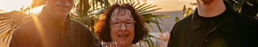 Three people are clinking wine glasses, surrounded by palm trees at sunset., © Weingut Bayer Three people are clinking wine glasses, surrounded by palm trees at sunset., © Weingut Bayer