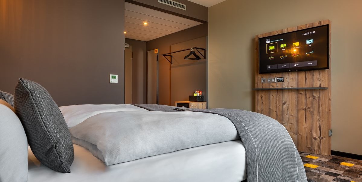 Modernes Hotelzimmer mit Doppelbett, grauen Kissen, Wand-TV und offener Garderobe. Der Boden hat ein gemustertes Design., © Novum Hospitality