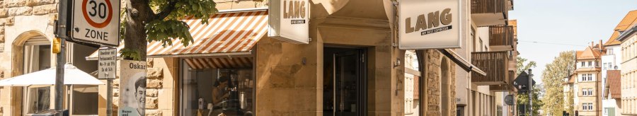 Caf&eacute; Lang an einer Stra&szlig;enecke mit Menschen, die drau&szlig;en sitzen. Ein 30er-Zone-Schild und B&auml;ume sind ebenfalls zu sehen., &copy; SMG, Sarah Schmid