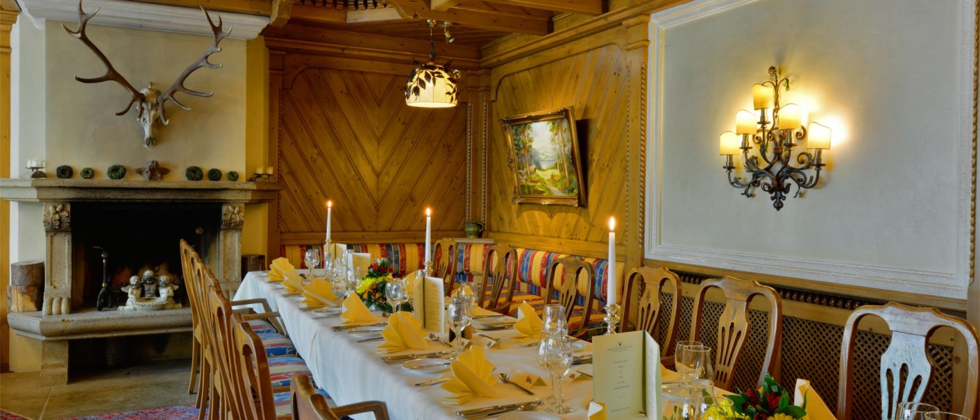 Elegant dining room with long, laid table, fireplace with antlers, wooden walls and paintings. Candles and flower arrangements decorate the room., © TOMAS