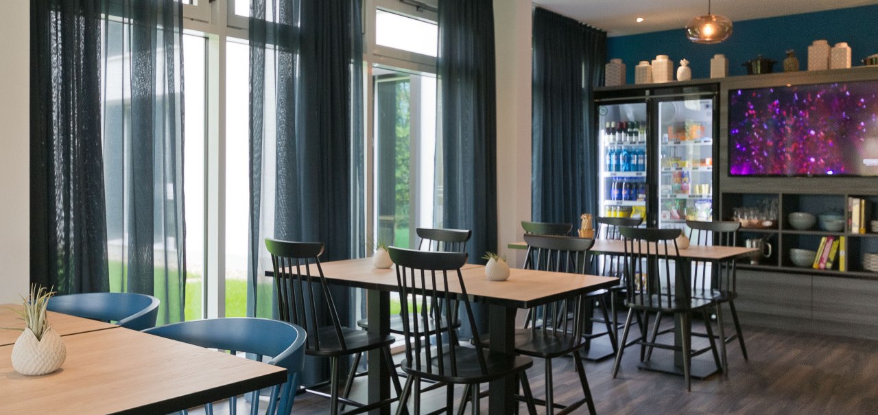 Moderner Raum mit Holztischen, schwarzen und blauen St&uuml;hlen. K&uuml;hlschrank mit Getr&auml;nken, Fernseher an der Wand. Dunkle Vorh&auml;nge und Holzboden., &copy; Arthotel Ana / elaya