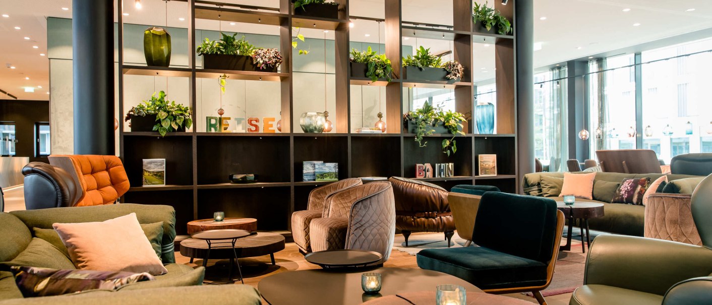Moderne Lobby mit bequemen Sesseln, Sofas und Tischen. Regale mit Pflanzen und Dekorationen, helle Beleuchtung und große Fenster., © Motel One - Stuttgart Hauptbahnhof