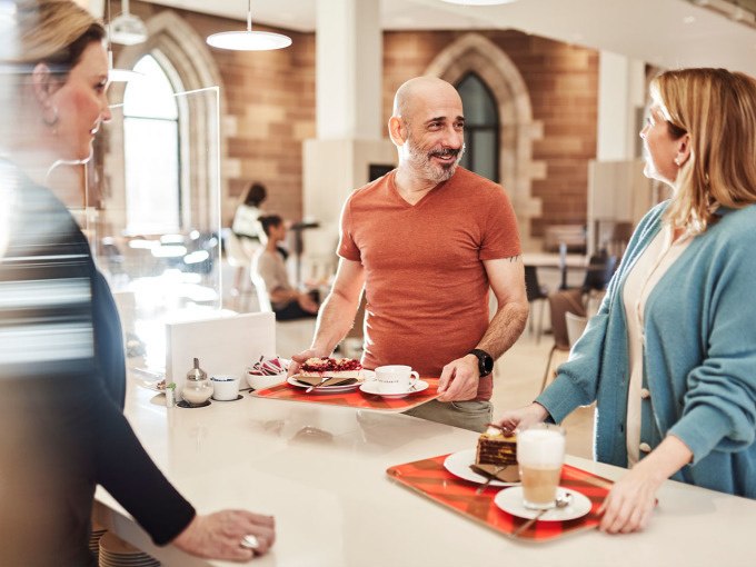 Drei Personen stehen in einem Café mit Tabletts voller Kaffee und Kuchen und unterhalten sich. Im Hintergrund sind große Fenster zu sehen., © Landesmuseum Württemberg Drei Personen stehen in einem Café mit Tabletts voller Kaffee und Kuchen und unterhalten sich. Im Hintergrund sind große Fenster zu sehen., © Landesmuseum Württemberg