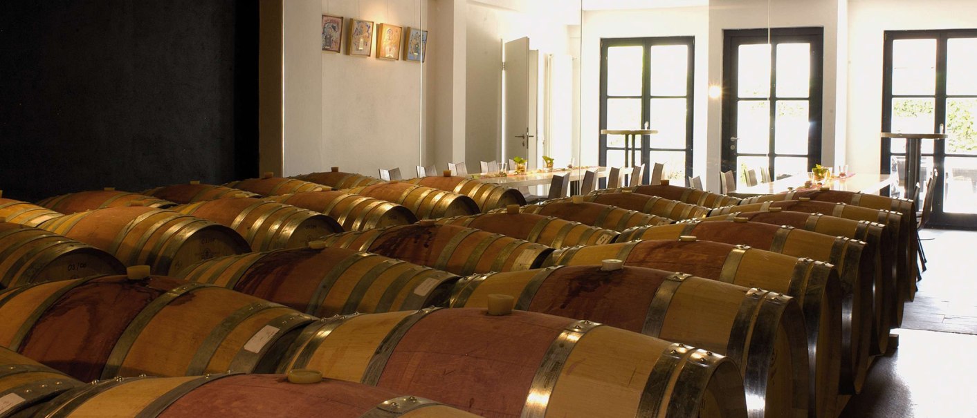 Ein Weinkeller mit vielen Holzfässern. Im Hintergrund steht ein langer Tisch mit Stühlen, dekoriert für eine Weinprobe., © Wöhrwag