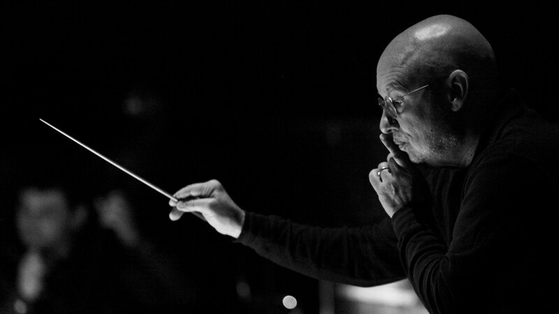 Ein Dirigent in Schwarz-Weiß, der mit einem Taktstock konzentriert ein Orchester leitet., © Württembergische Staatstheater Stuttgart Ein Dirigent in Schwarz-Weiß, der mit einem Taktstock konzentriert ein Orchester leitet., © Württembergische Staatstheater Stuttgart
