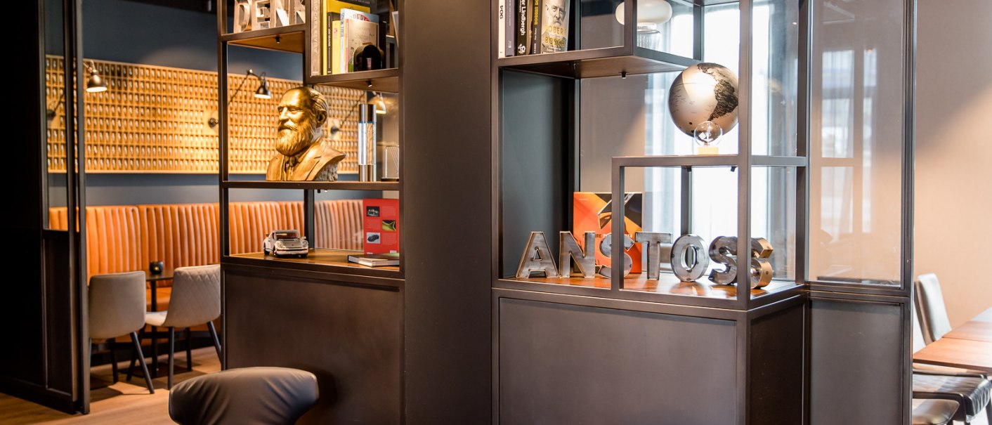 Modernes Café mit Regalen, Büchern, einer Büste und einem Globus. Sitzbereich mit braunen Polstern und stilvollen Stühlen., © TOMAS