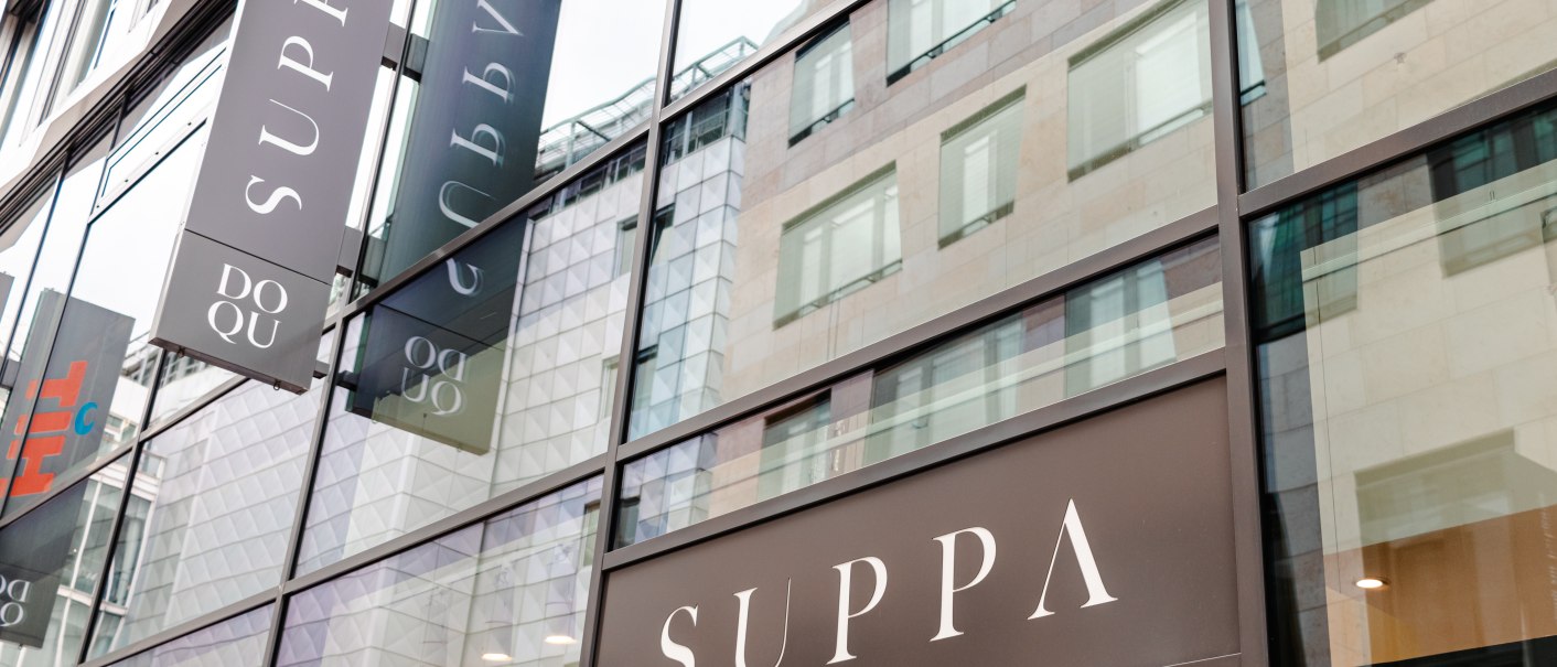 Glasfassade eines modernen Gebäudes mit mehreren SUPPA-Schildern. Die Spiegelung zeigt umliegende Architektur., © SMG_Sarah Schmid Glasfassade eines modernen Gebäudes mit mehreren SUPPA-Schildern. Die Spiegelung zeigt umliegende Architektur., © SMG_Sarah Schmid