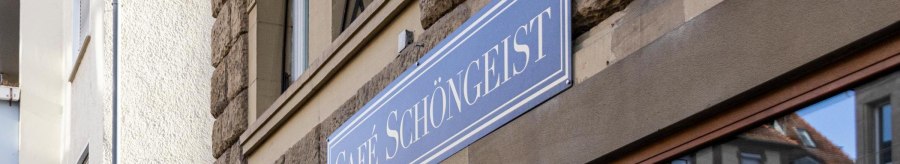 Das Bild zeigt das Schild des Caf&eacute;s Sch&ouml;ngeist an einer steinernen Hausfassade. Im Fenster spiegelt sich ein gegen&uuml;berliegendes Geb&auml;ude., &copy; Stuttgart-Marketing GmbH, Sarah Schmid