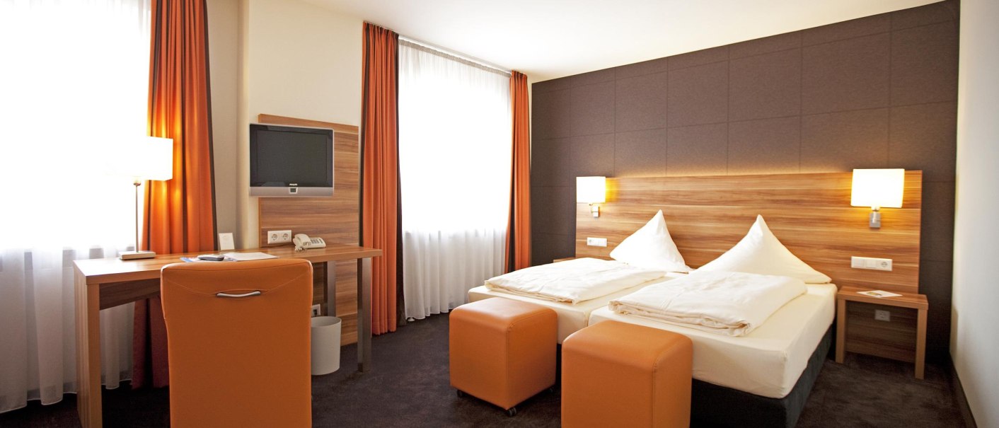 Modernes Hotelzimmer mit zwei Einzelbetten, orangefarbenen Vorhängen, Schreibtisch, Fernseher und zwei Hockern. Helle und gemütliche Atmosphäre., © Hotel Hirsch