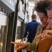 Zwei Frauen stehen vor einem Weinspender und zeigen auf verschiedene Weinflaschen. Im Hintergrund sind weitere Flaschen und Gläser zu sehen., © Stuttgart Marketing GmbH, Fotografin Martina Denker Zwei Frauen stehen vor einem Weinspender und zeigen auf verschiedene Weinflaschen. Im Hintergrund sind weitere Flaschen und Gläser zu sehen., © Stuttgart Marketing GmbH, Fotografin Martina Denker