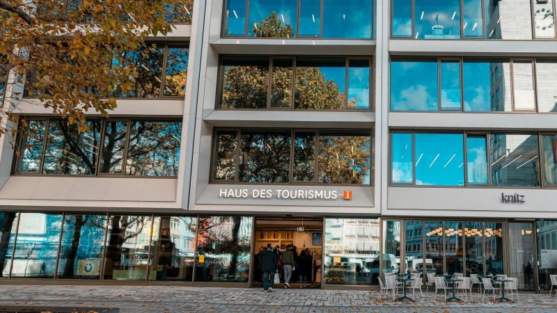 Moderne Glasfassade des Haus des Tourismus mit Spiegelungen von B&auml;umen und blauem Himmel. Menschen betreten die Tourist-Information im Geb&auml;ude, drau&szlig;en stehen Tische und St&uuml;hle., &copy; SMG, Sarah Schmid