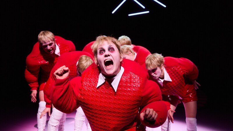 Theaterszene mit mehreren Personen in roten, gepolsterten Kostümen und weißem Make-up. Eine Person im Vordergrund schreit, im Hintergrund leuchten Neonlichter., © Württembergische Staatstheater Stuttgart Theaterszene mit mehreren Personen in roten, gepolsterten Kostümen und weißem Make-up. Eine Person im Vordergrund schreit, im Hintergrund leuchten Neonlichter., © Württembergische Staatstheater Stuttgart